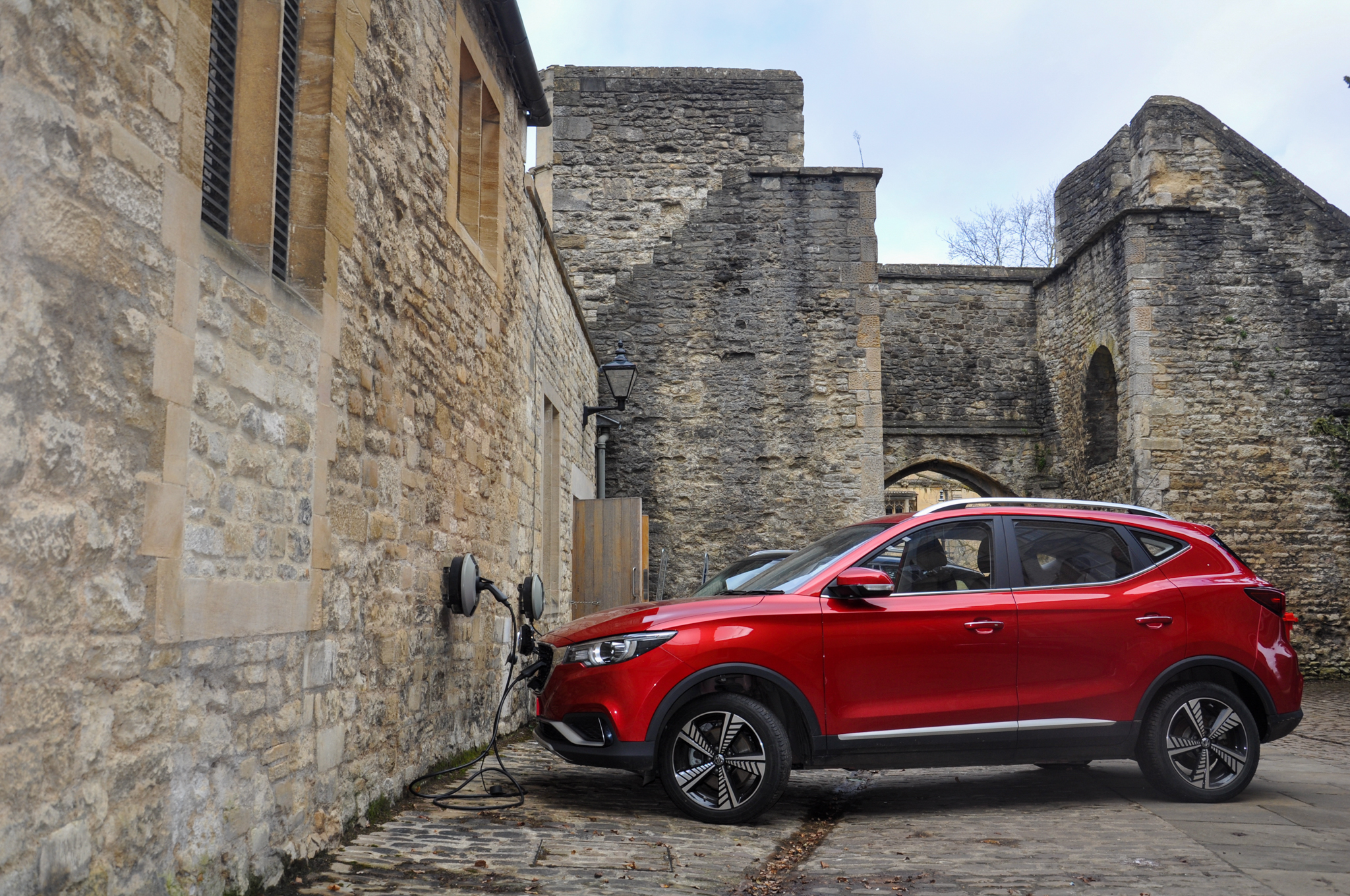 A red car being charged at an electric vehicle charging point, with old buildings and medieval city wall in the background