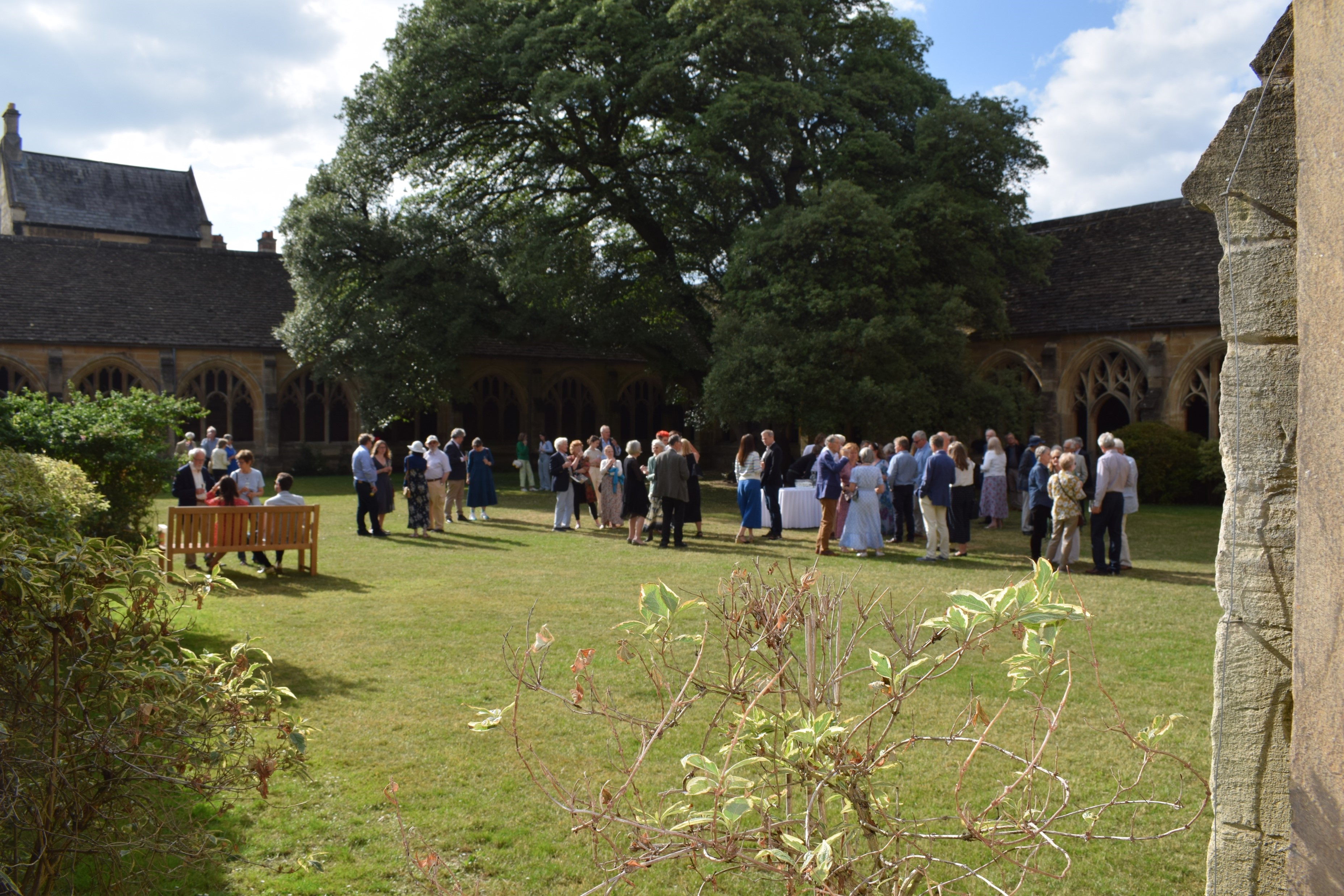 Large group of people on central lawn of some medieval cloisters, with a large oak tree behind