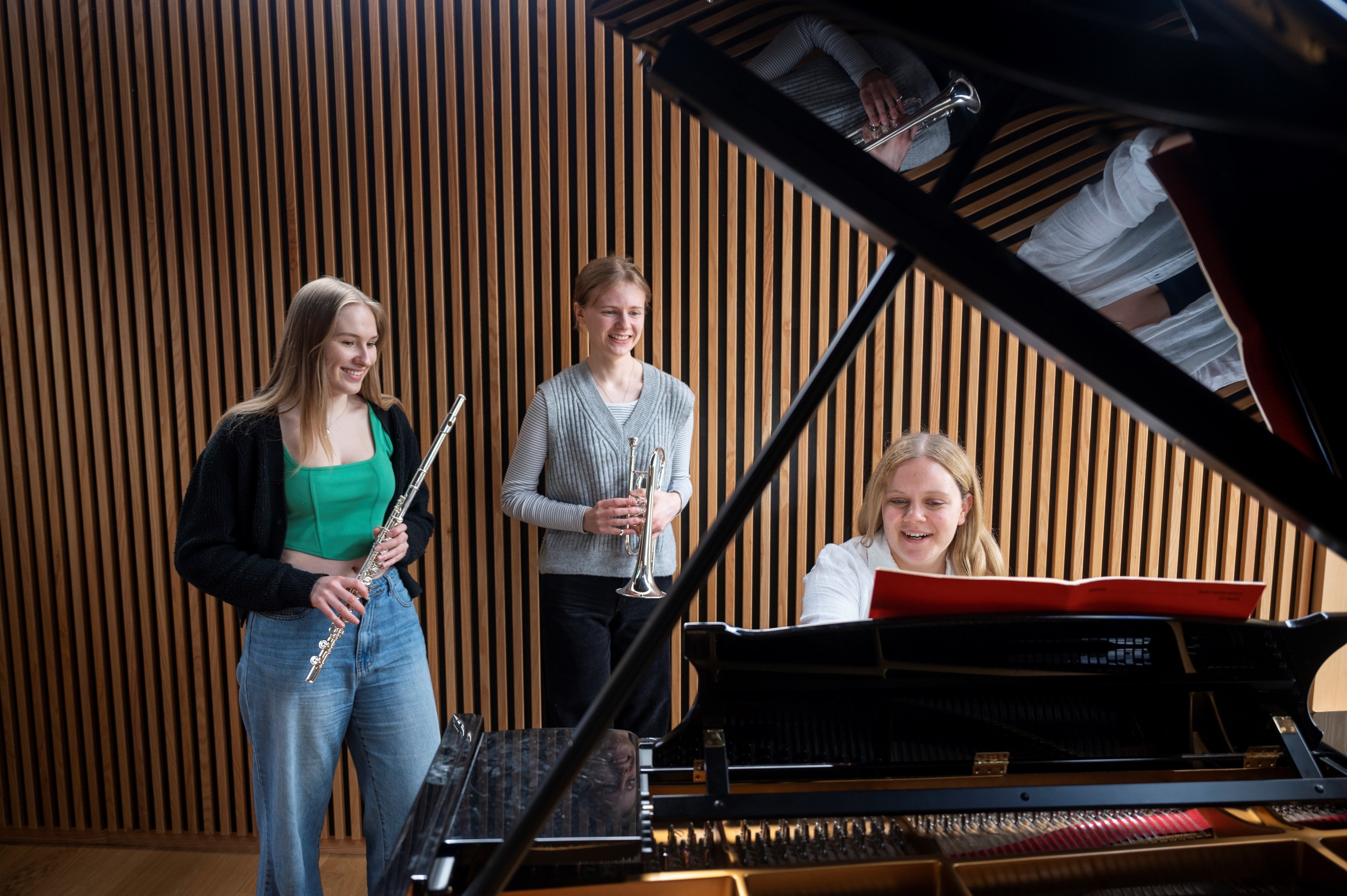 A student plays the piano whilst two other students hold instruments (a flute and a cornet)