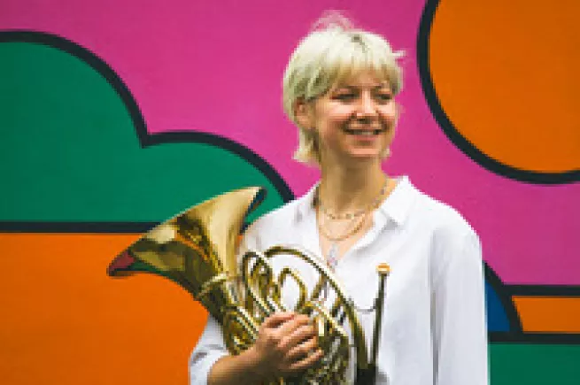 Blonde-haired woman in white shirt stands in front of a coloured wall holding a french horn.