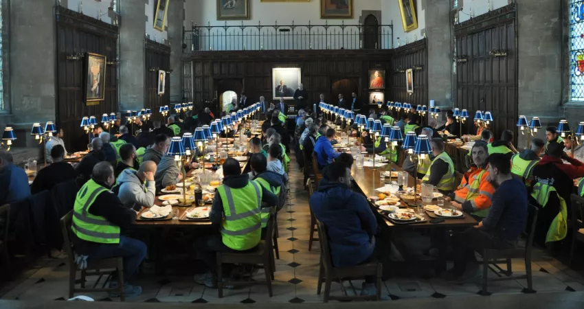 Warden standing on a chair giving a speech to hundreds of people in high vis clothing eating breakfast; all in a medieval Dining Hall