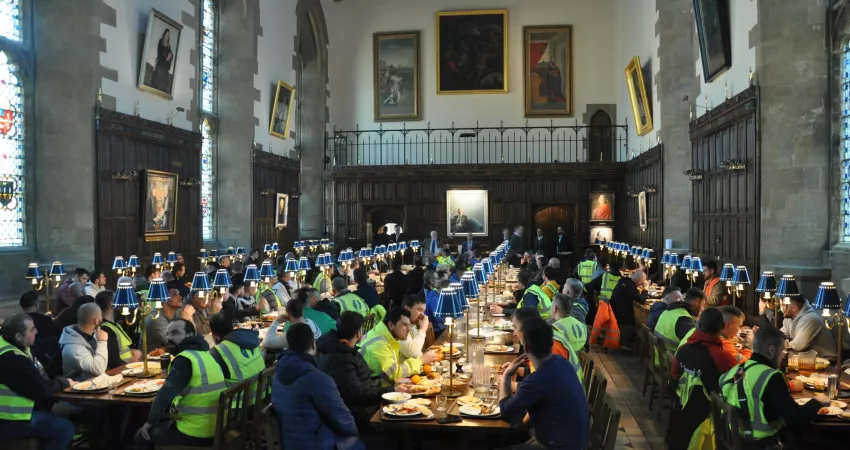 Bursar giving a speech to hundreds of people in high vis clothing eating breakfast; all in a medieval Dining Hall