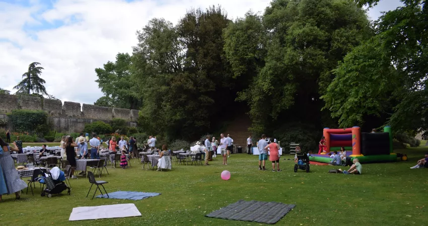 People sat around tables on a large lawn with a Mound covered in trees behind