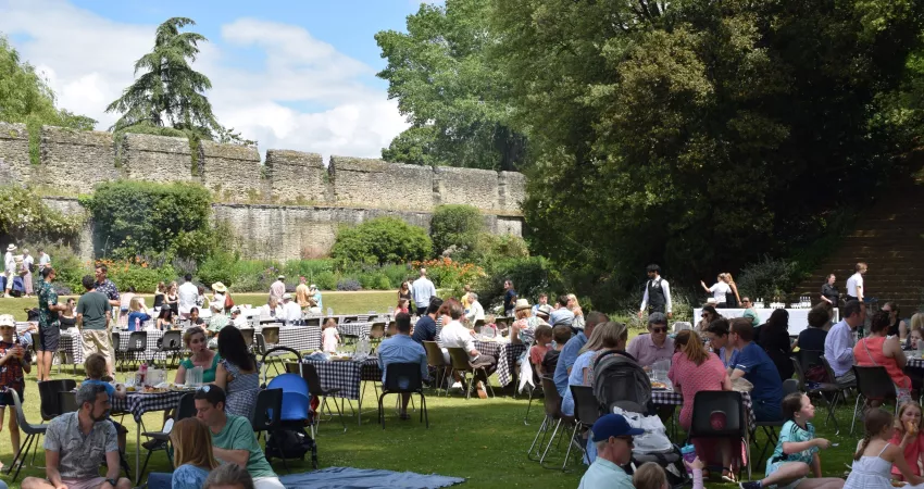 People sat on chairs in small groups with trees and a medieval city wall behind