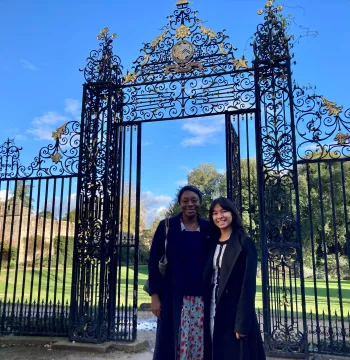 Grace and Ei standing in front of an elaborate iron screen in Garden Quad, with city wall behind