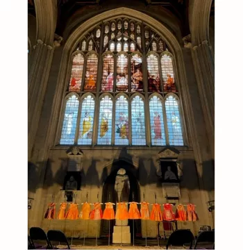 Thirteen orange dresses lined up in a row underneath an ornate stained glass, arched window
