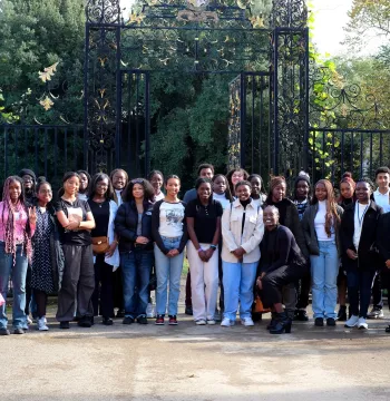 A large group of Black British students standing smiling at the camera in front of an ornate iron screen