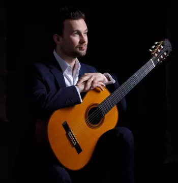 Man sits facing side-on to the camera with his hands placed on top of a classical guitar. Black background