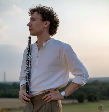 Man stands (facing side-on to the camera) in field, holding oboe.