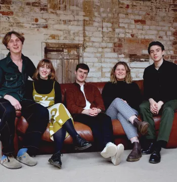 A row of five people sit on a sofa in an empty space with a brick wall behind them.